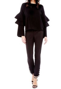 Schwarzer Pullover mit schichtigen Rüschensäumen, kombiniert mit schwarzen Hosen mit Knopfakzenten am Saum. Abgerundet mit schwarzen Riemchenheels.