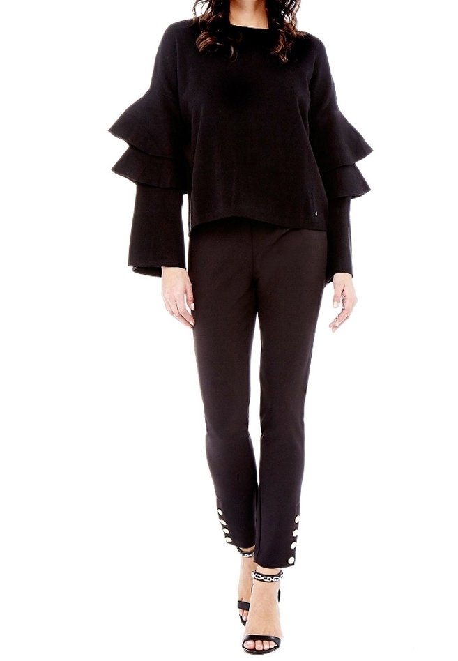 Schwarzer Pullover mit schichtigen Rüschensäumen, kombiniert mit schwarzen Hosen mit Knopfakzenten am Saum. Abgerundet mit schwarzen Riemchenheels.