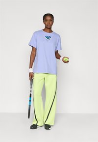 Μπλουζάκι λεβάντας με γραφικό ρακέτας τένις, συνδυασμένο με φαρδιά παντελόνια νέον πράσινου χρώματος με μαύρες ρίγες στο πλάι, κρατώντας μπάλα και ρακέτα τένις.