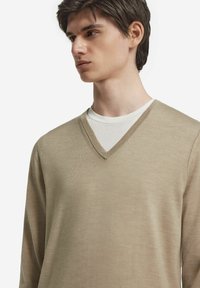 Beige V-ringad tröja i lättviktsmaterial med en texturerad yta. Lager på en ljusfärgad rundhalsad skjorta med en ren halsringning.