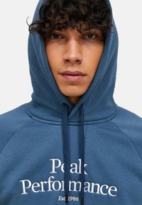 Blauwe hoodie gemaakt van zacht materiaal met een trekkoordcapuchon. Bevat witte tekst "Peak Performance Est. 1986" op de borst en raglanmouwen.
