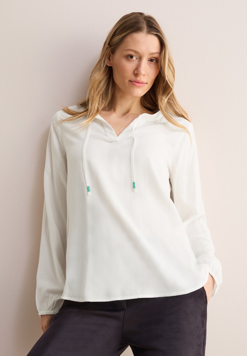 Witte blouse met lange mouwen, een V-hals en een trekkoordkap, gemaakt van lichte stof en met twee groene toggles aan de uiteinden van de trekkoorden.