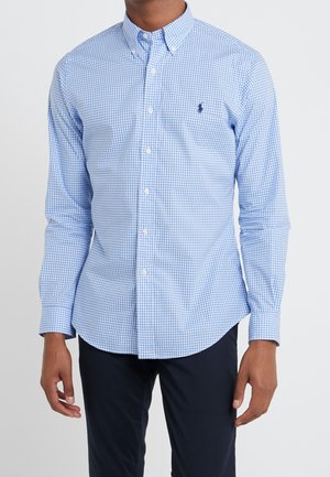 Man wearing a blue and white checkered long-sleeve button-up shirt with black pants, standing against a plain background.