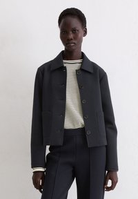 Czarna krótka kurtka zapinana na guziki z dwoma przednimi kieszeniami, noszona na paskowaną koszulę z długim rękawem, w zestawie z dopasowanymi ciemnymi spodniami.