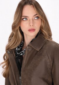 Giacca di pelle marrone con una texture liscia, dotata di chiusura con zip e colletto classico. Una sciarpa a motivi bianchi e neri aggiunge un tocco di dettagli.
