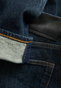 Tume sinine teksapüksid helesinise mansetiga, oranži õmblusega ja musta nahast plaastriga, millel on reljeeftekst.