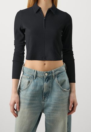 Vrouw draagt een zwart geribd cropped top met lange mouwen en ritssluiting en lichtblauwe hoge taille jeans tegen een effen achtergrond.