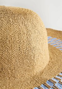 Strohoed met een brede rand, beige van kleur, met een geweven textuur en franjes. Heeft een lichtblauwe accent nabij de rand.