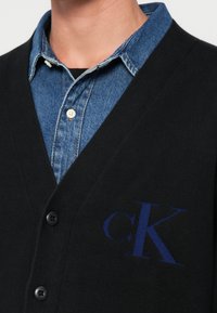 Cardigã preto com um design em forma de V, apresentando um logótipo azul "CK" bordado no peito, combinado com uma camisa de ganga abotoada por baixo.