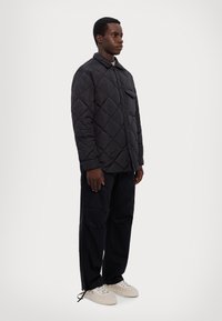 Giacca nera imbottita e trapuntata con design colletto, tasca frontale e bottoni a pressione, abbinata a pantaloni cargo neri e sneakers beige.