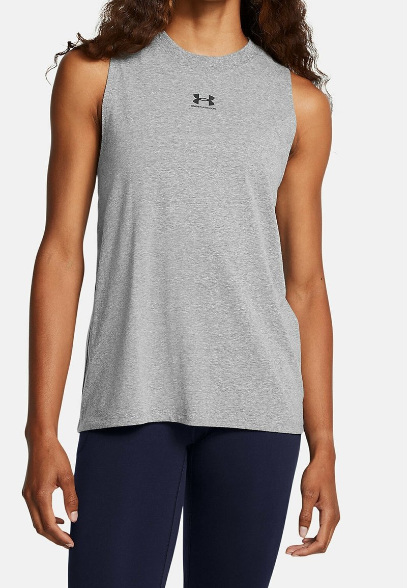 Haut du corps d'une personne portant un débardeur gris clair Under Armour et un pantalon bleu foncé sur un fond uni.