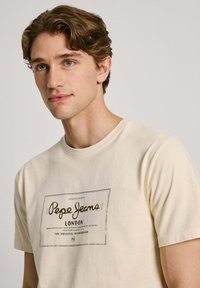 Kremo spalvos medvilnės marškinėliai su juodu spaudos logotipu, kuriame užrašyta "Pepe Jeans London", trumpomis rankovėmis ir klasikine apvalia iškirpte.