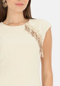 Beżowa sukienka z fakturowanym koronkowym ramieniem, zdobiona kaskadowymi frędzlami. Gładki materiał o dopasowanym kroju i okrągłym dekolcie.