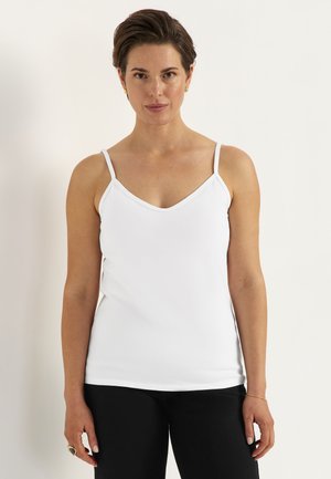 Frau mit kurzem braunem Haar, die ein weißes ärmelloses V-Ausschnitt-Top und schwarze Hose trägt, stehend vor einem schlichten hellen Hintergrund.