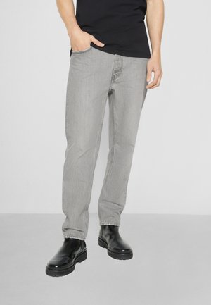 Person wearing light gray jeans, black boots, and a black shirt, with one hand in pocket against a plain light background.