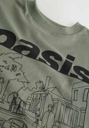 Zelené tričko s čiernym obrysovým ilustráciou budov a ľudí, s nápisom "oáza" výrazne zobrazeným nad ním.
