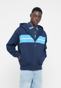 Marineblå zip-up hættetrøje med to vandrette lyseblå striber på brystet, en hætte med snor og sidelommer.