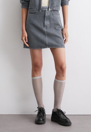 Vrouw met een grijze spijkerrok met krijtstrepen, doorzichtige beige kniekousen en zwarte platform veterpumps, staand tegen een witte muur.