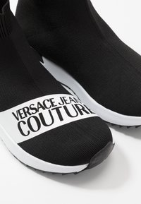 Musta neulotut slip-on-tennarit, joissa valkoinen logonauha, jossa lukee "VERSACE JEANS COUTURE." Karkea kangas ja kumipohja.
