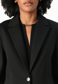 Zwarte blazer met een gelapte kraag, enkele zilveren knoopsluiting en een minimalistisch ontwerp. Zacht materiaal met een gladde textuur.