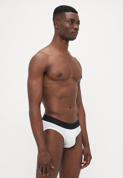 Hvide briefs med en sort elastisk talje, der har et logo. Stoffet ser glat ud med et tætsiddende design. Model står sidelæns.