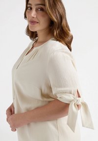 Cremfarbene Bluse mit gepufften Ärmeln, Bindebändern und einem strukturierten Stoff. Knopfleiste vorne mit kleinen Falten am Ausschnitt.