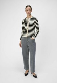  gestreifter Cardigan in Schwarz und Weiß mit Rundhalsausschnitt und Knopfleiste, kombiniert mit lockeren grauen Jeans und spitzen schwarzen High Heels.