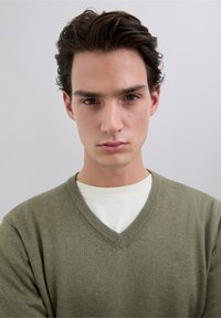 Suéter de punto color verde oliva con cuello en V y ribete acanalado, superpuesto sobre una camisa blanca. Presenta un detalle sutil del logo en el lado izquierdo.