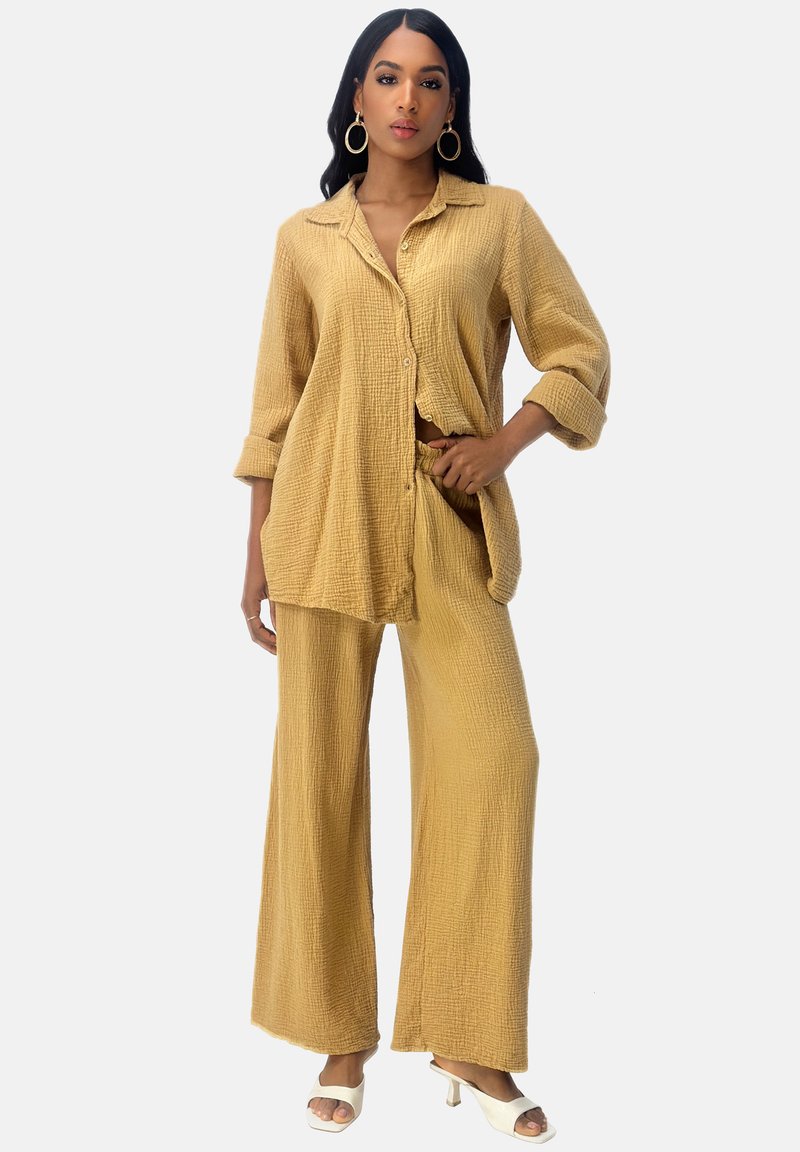 Camicia sovradimensionata di un giallo chiaro, con texture, bottoni e maniche arrotolate, abbinata a pantaloni larghi coordinati; il modello indossa tacchi bianchi.