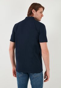 Námorně modrá polo košile s krátkým rukávem vyrobená z bavlny, která má klasický límec a jednoduchý design bez viditelných log a vzorů.