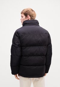 Black puffer jacket with a high collar, quilted design, and visible stitching. Features a relaxed fit and side pockets for functionality.