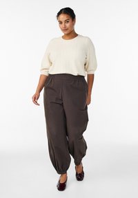 Suéter de punto beige claro con textura y mangas cortas, combinado con pantalones cónicos de corte ancho en marrón oscuro y zapatos tipo slip-on burdeos.