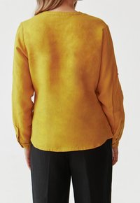 Camicia a maniche lunghe in lino giallo vivace con scollo rotondo, texture liscia e orlo curvo, abbinata a pantaloni neri.