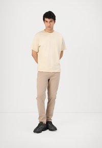 Beige kortermet t-skjorte kombinert med lysebrune bukser og svarte joggesko. Enkelt design uten synlige mønstre eller aksenter.