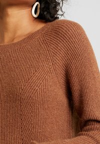 Brauner Strickpullover mit gerippter Textur, weitem Rundhalsausschnitt und betonten Nähten für eine taillierte Passform.