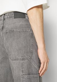 Pantalones cargo de mezclilla gris con un acabado texturizado, que cuentan con un gran bolsillo lateral y una etiqueta rectangular negra en la cintura.