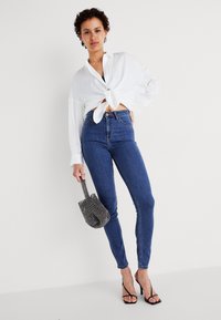 Vit knytblus, högmidjade mörkblå skinny jeans, svarta klackar med remmar och en texturerad svart och vit väska. Enkel, strömlinjeformad design.