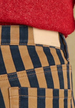 Dryžuotos kelnės tamsiai mėlynos ir rudos spalvos, pagamintos iš džinso, su rudos odos lopeliu ant liemens, pažymėtu užrašu „JOULES“. Matomi tekstūruoti siūlės.