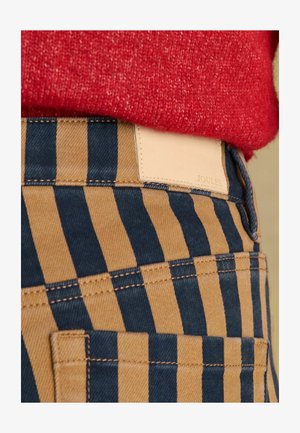 Spodnie w paski w kolorach granatowym i beżowym, wykonane z denimu, z beżową skórzaną naszywką z napisem "JOULES" na pasku. Widoczne fakturowane przeszycia.