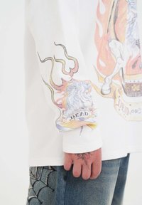 Witte textuur lange mouw shirt met kleurrijke vlammende leeuw print en "DEAD" tekst op de mouw, gedragen met blauwe spijkerbroek met spinnenweb borduursel.