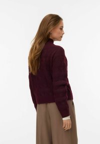 Bordowy dzianinowy sweter z wysokim kołnierzem, teksturowanym wzorem i prążkowanymi mankietami. Nosi się go z jasnymi spodniami; tło jest jednolicie białe.