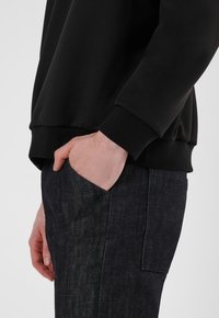 Schwarzer Sweatshirt mit glatter Textur, ausgestattet mit einem gerippten Saum und Bündchen, kombiniert mit dunklen Jeanshosen mit sichtbaren Nähten und Seitentaschen.