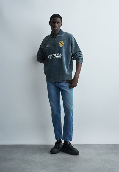 Άντρας στέκεται μπροστά σε απλό τοίχο φορώντας σκούρο γκρι φούτερ με φερμουάρ στο λαιμό και λογότυπα, μπλε τζιν και μαύρα αθλητικά παπούτσια, με τα χέρια στις τσέπες.