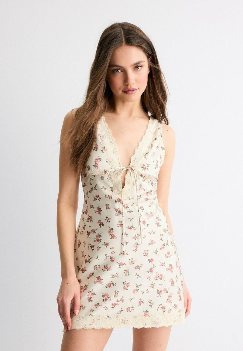Jeune femme aux longs cheveux portant une robe sans manches crème à fleurs avec bordure en dentelle et lien à l'avant, debout devant un fond blanc uni.