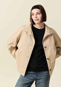 Beżowa, oversize'owa kurtka z dużym kołnierzem, matową fakturą i czarnymi guzikami. W zestawie z czarnym swetrem i dżinsowymi spodniami.