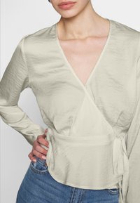 Blusa avvolgente beige chiaro con maniche lunghe, caratterizzata da un tessuto testurizzato, scollatura a V e dettaglio con fiocco laterale, abbinata a jeans denim blu.