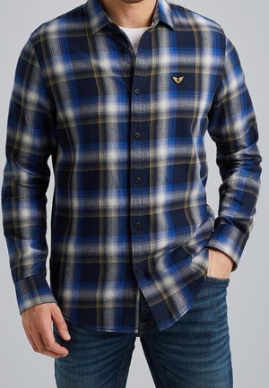 Homme portant une chemise à manches longues à carreaux bleu, blanc et noir avec un petit emblème jaune sur la poitrine, associée à un jean bleu.