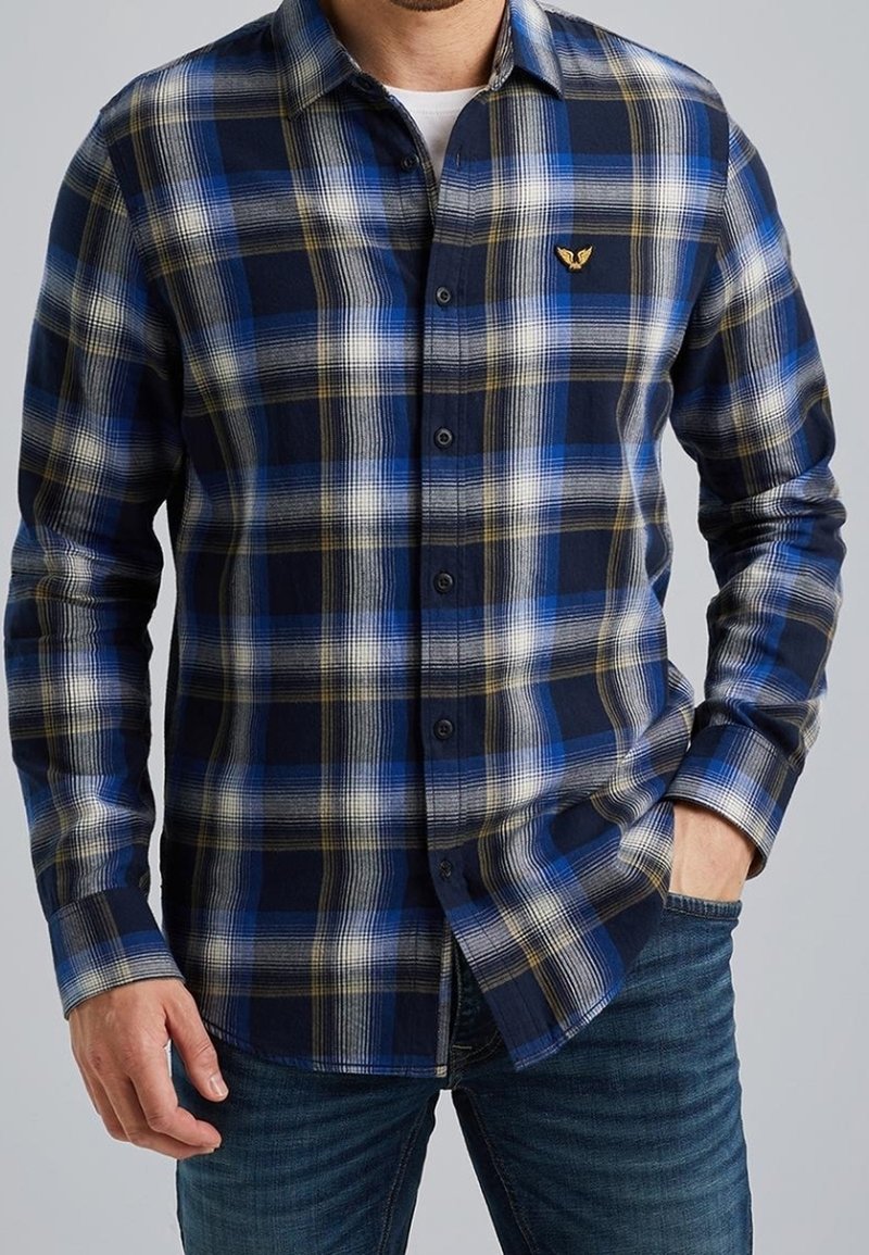 Homme portant une chemise à manches longues à carreaux bleu, blanc et noir avec un petit emblème jaune sur la poitrine, associée à un jean bleu.