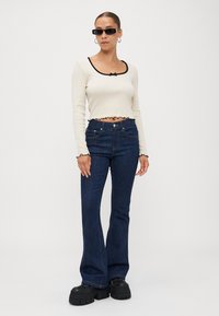 Cream cropped top with black trim and ruffle details, paired with dark blue flared jeans. Model wears chunky black platform shoes.