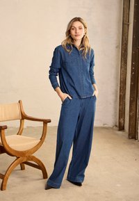 Femme debout les mains dans les poches, portant une chemise en jean bleue assortie et un pantalon large près d'une chaise en bois dans un cadre intérieur neutre.